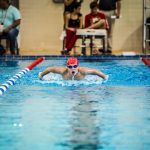 A young swimmer in a red cap competes in a butterfly stroke during an indoor pool race.